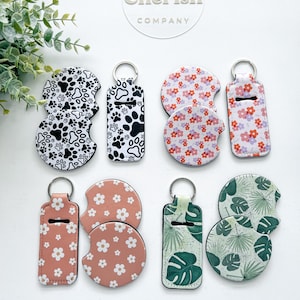 May include: A collection of car coasters and keychains with various designs. The coasters feature paw prints, floral patterns, and tropical leaves. The keychains have matching designs. The items are in black, white, pink, and green.