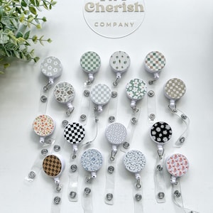 May include: A collection of fifteen circular badge reels with retractable clear plastic straps, featuring various patterns including checkered, floral, and botanical designs. A circular acrylic sign with the text Two Cherish COMPANY is visible.