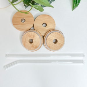 May include: A set of four bamboo lids with a hole and two clear glass straws. The lids have a natural wood grain finish and a silicone seal. The straws are straight and bent. The items are arranged on a white surface.
