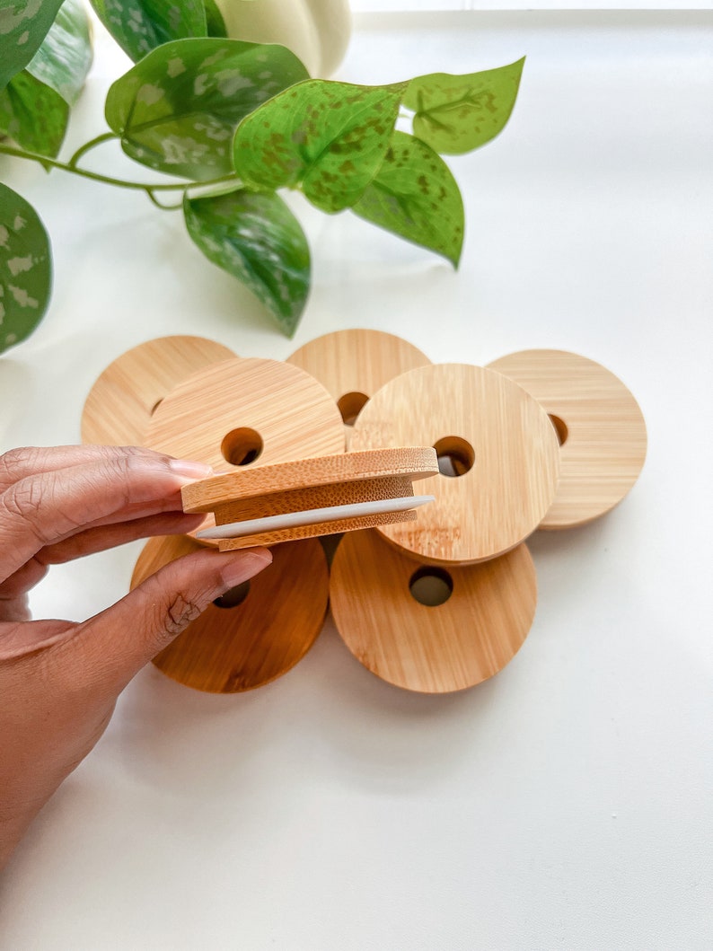 May include: A collection of round bamboo lids with a central hole, some stacked, others scattered. One lid is held up, revealing a white silicone seal. The lids are light brown, with a natural wood grain. The background is white.