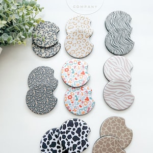 May include: A collection of car coasters with various animal print designs, including leopard, zebra, cow, and giraffe patterns. The coasters are in neutral tones of beige, gray, and brown, with a black border. The brand name "Two Cherish Company" is visible in the background.