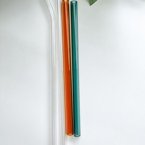May include: Four glass straws in clear, orange, and teal. Two are straight, one is bent, and all have a smooth, cylindrical design. The straws are arranged vertically against a white background, with a green leaf in the upper right corner.