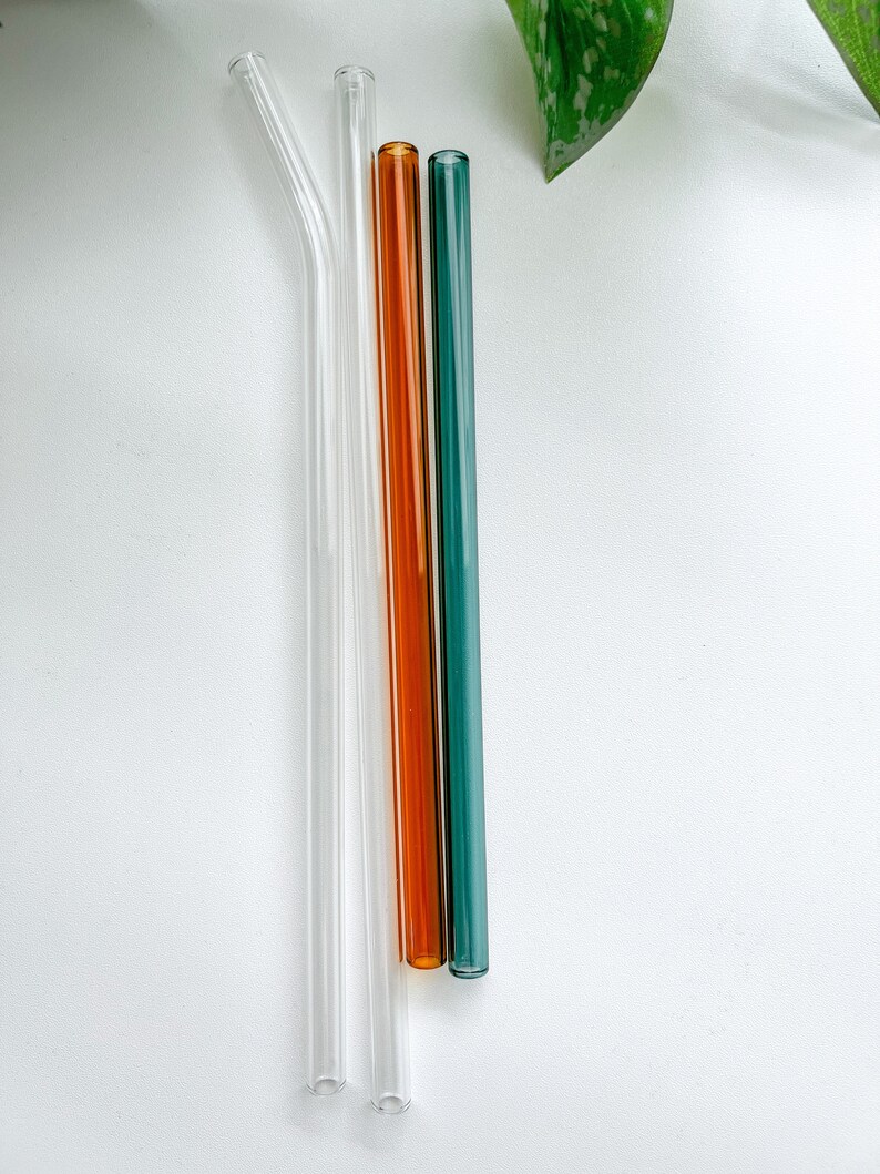 May include: Four glass straws in clear, orange, and teal. Two are straight, one is bent, and all have a smooth, cylindrical design. The straws are arranged vertically against a white background, with a green leaf in the upper right corner.