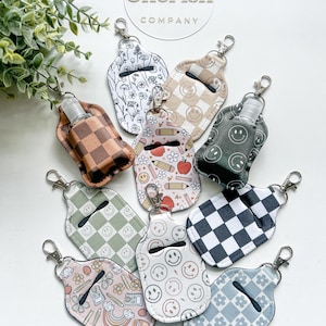 May include: A selection of hand sanitiser holders with diverse patterns, such as smiley faces, floral, and chequered designs. Each holder features a silver clip and a small hand sanitiser bottle. The brand name "Two Cherish Company" is visible.