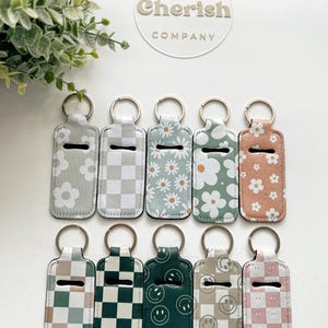 K&ouml;nnte beinhalten: Eine Sammlung von zehn Schl&uuml;sselanh&auml;ngern mit verschiedenen Mustern, darunter G&auml;nsebl&uuml;mchen, Schachbrettmuster und Smiley-Gesichter. Die Schl&uuml;sselanh&auml;nger sind in Gr&uuml;n-, Rosa- und neutralen Farbt&ouml;nen gehalten. Jeder Schl&uuml;sselanh&auml;nger hat einen silbernen Ring und einen schwarzen Schlitz.