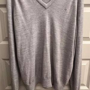 Può includere: Un maglione grigio a collo a V a maniche lunghe con un piccolo logo sul petto sinistro. Il maglione è realizzato in un materiale morbido e confortevole.