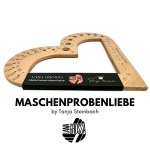 Könnte beinhalten: Ein herzförmiger Holzmaßstab mit Markierungen zum Messen von Strickmaschen und -reihen. Der Maßstab ist mit "LANA GROSSA Maschenprobenliebe" und "Tanja Steinbach" beschriftet.