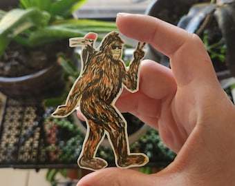 May include: A Bigfoot sticker with a peace sign and sunglasses, accompanied by a woodpecker. The sticker features brown, orange, and black colors, with a white border. The Bigfoot is depicted walking.