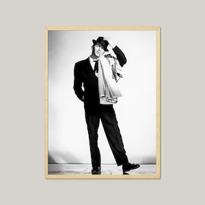 May include: Black and white photo of a man wearing a suit and a hat. He is standing with his right hand on his hip and his left hand holding his hat. He is looking to the side.
