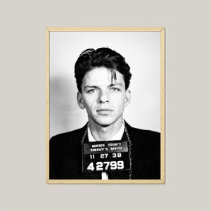 May include: Black and white framed portrait of a man in a suit, with dark hair and a serious expression. A sign around his neck reads "Bergen County Sheriff's Office" with numbers. The image is a vintage style photograph.