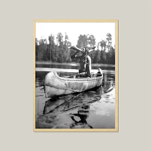 May include: A black and white photograph of a Native American man in a canoe, paddling with a wooden paddle. He is wearing traditional clothing and a feathered headdress. The canoe is on a calm lake with trees in the background.