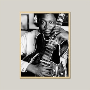 May include: Black and white framed photograph of a man holding a guitar. The guitar is a dark color with a white fretboard. The man is wearing a light-colored shirt and a ring on his finger.