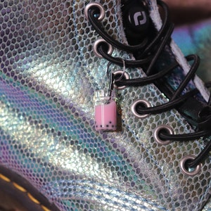 Puede incluir: Un par de botas iridiscentes con estampado de piel de serpiente, cordones negros y un charm de té de boba rosa y negro atado a los cordones.