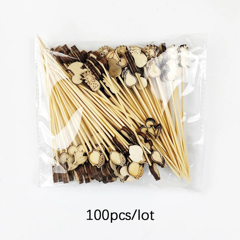 Może przedstawiać: Przezroczysty plastikowy worek wypełniony 100 drewnianymi szaszłykami z r&oacute;żnymi kształtami owoc&oacute;w i warzyw na końcach. Tekst "100pcs/lot" jest nadrukowany pod workiem.