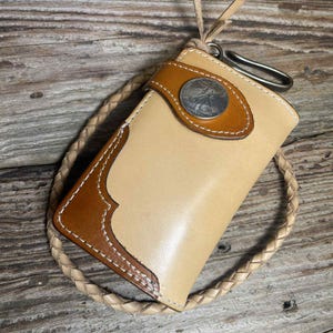 May include: A tan leather wallet with brown accents and white stitching. The wallet features a silver coin detail and a braided leather strap. A metal clasp is attached to the wallet.