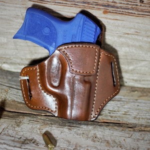 May include: A brown leather holster with a blue handgun inside. The holster has stitching around the edges and a belt loop. There are three bullet casings on the wooden surface.