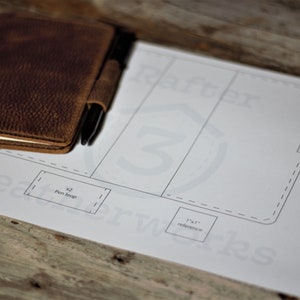 May include: A brown leather notebook cover with a pen loop and a pen inside. The cover is laying on a white paper pattern with the text "Leatherworks" and a 1x1 inch reference square.