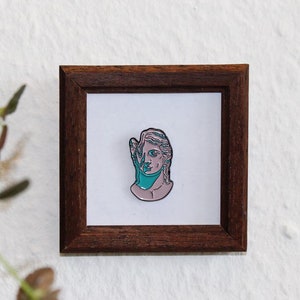 Könnte beinhalten: Eine gerahmte Emaille-Anstecknadel mit einem Frauenbüstenbild mit einem türkisfarbenen Schal. Die Anstecknadel befindet sich in einem braunen Holzrahmen.