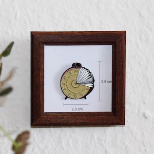 Könnte beinhalten: Gerahmter, goldfarbener Uhren-Ansteckpin mit einem buchähnlichen Design, 2,8 cm hoch und 2,3 cm breit. Der Pin befindet sich vor einem weißen Hintergrund in einem dunkelbraunen Holzrahmen.