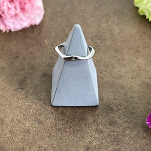 Könnte beinhalten: Ein silberner Ring mit wellenförmigem Design wird auf einem grauen, geometrischen Ständer präsentiert. Die Oberfläche des Rings reflektiert das Licht. Rosa und gelbe Blumen sind im Hintergrund auf einer braunen Oberfläche.