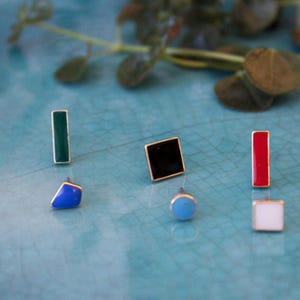 Könnte beinhalten: Eine Sammlung bunter Ohrstecker mit goldfarbenen Rändern. Die Ohrringe haben verschiedene Formen, darunter Rechtecke, Quadrate und Kreise, in Grün, Schwarz, Rot, Blau, Hellblau und Weiß. Sie sind auf einer türkisfarbenen Oberfläche angeordnet.