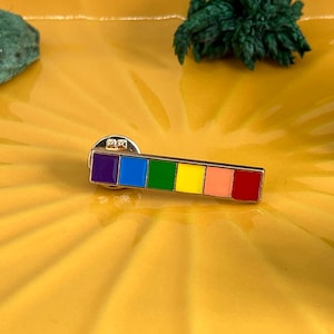 Peut inclure: Une épingle rectangulaire en émail aux couleurs de l'arc-en-ciel, avec du violet, du bleu, du vert, du jaune, de l'orange et du rouge. L'épingle a une bordure dorée et un fermoir. L'épingle est sur une surface jaune.