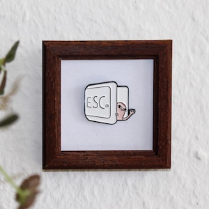 Könnte beinhalten: Eine weiße Emaille-Anstecknadel mit einer Comicfigur, die aus einer "ESC"-Taste schaut. Die Anstecknadel ist in einem braunen Holzrahmen ausgestellt.