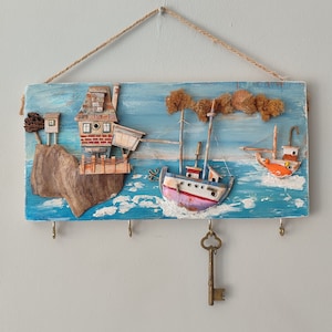 Puede incluir: Un porta llaves de madera con temática náutica. Presenta un barco azul y blanco, una pequeña casa en un acantilado y una llave colgando de un gancho. El fondo está pintado para parecerse al océano.