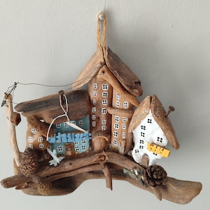 Puede incluir: Una escultura de madera de un pequeño pueblo con tres casas, un árbol y un pájaro. Las casas están pintadas de blanco con ventanas y puertas negras. El árbol está hecho de madera flotante y tiene un pájaro posado en una rama. La escultura está montada en un trozo de madera flotante.