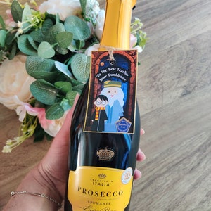 May include: A bottle of Prosecco with a yellow label that says "Prosecco Spumante Extra Dry" and a gold crown. The bottle has a tag attached to it that says "To the Best Teacher Since Dumbledore" with a cartoon image of Harry Potter and Dumbledore.