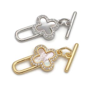 Può includere: Due chiusure a scatto in tono argento e oro con un design a quadrifoglio. Il quadrifoglio è realizzato in madreperla bianca e ha strass trasparenti attorno al bordo.