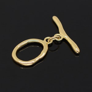 May include: Gold-toned metal toggle clasp with a smooth, rounded oval ring and a straight bar. The clasp is designed for attaching jewelry or other accessories.