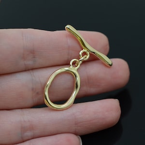 May include: Gold-toned metal toggle clasp with an oval ring. The clasp is designed with a smooth, organic shape.