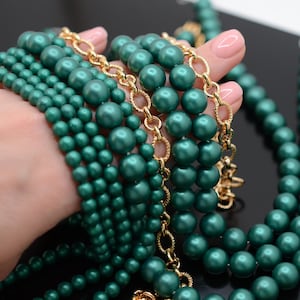 May include: A close-up of a hand holding a stack of emerald green pearl necklaces with a gold chain interwoven. The necklaces are made of different sized pearls and are arranged in a cascading pattern.
