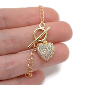 Può includere: Un braccialetto dorato con un ciondolo a forma di cuore. Il ciondolo è ricoperto di pietre di zircone cubico trasparenti. Il braccialetto ha una chiusura a scorrimento.
