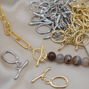 May include: A gold chain necklace with a toggle clasp and brown agate beads. The necklace is on a light brown fabric background. There are also silver jump rings in the background.