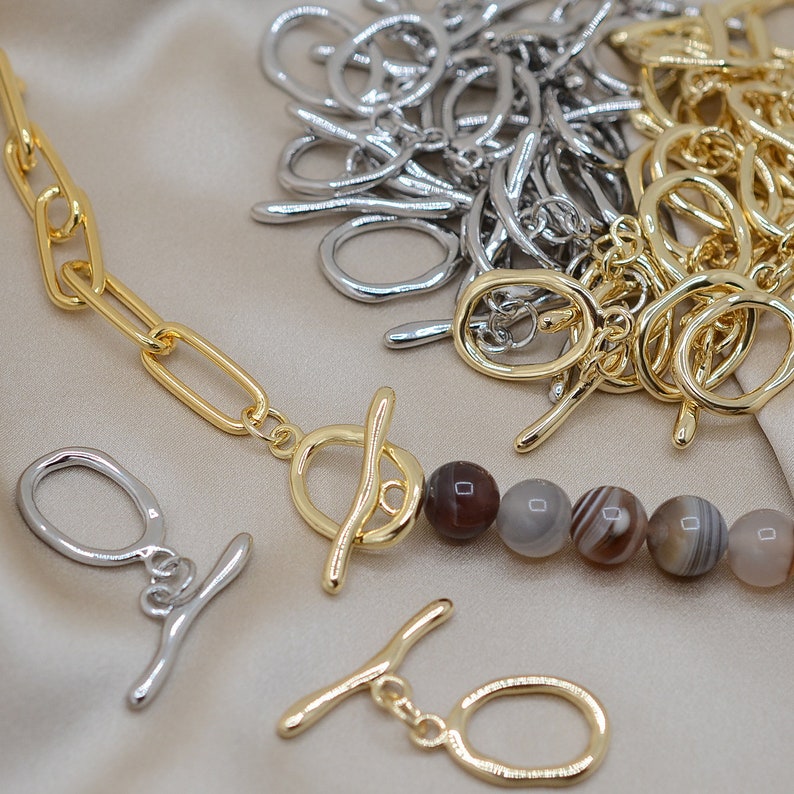 May include: A gold chain necklace with a toggle clasp and brown agate beads. The necklace is on a light brown fabric background. There are also silver jump rings in the background.