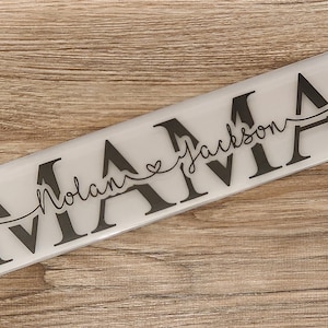 Puede incluir: Un letrero rectangular blanco con la palabra "MAMA" en letras negras grandes. Las palabras "Nolan" con un símbolo de corazón y "Jackson" están escritas en cursiva debajo de la palabra "MAMA".