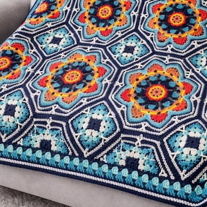May include: A handmade crochet blanket with a geometric pattern. The blanket features a navy blue background with colorful floral motifs in shades of red, orange, yellow, and blue. The blanket is resting on a gray sofa.