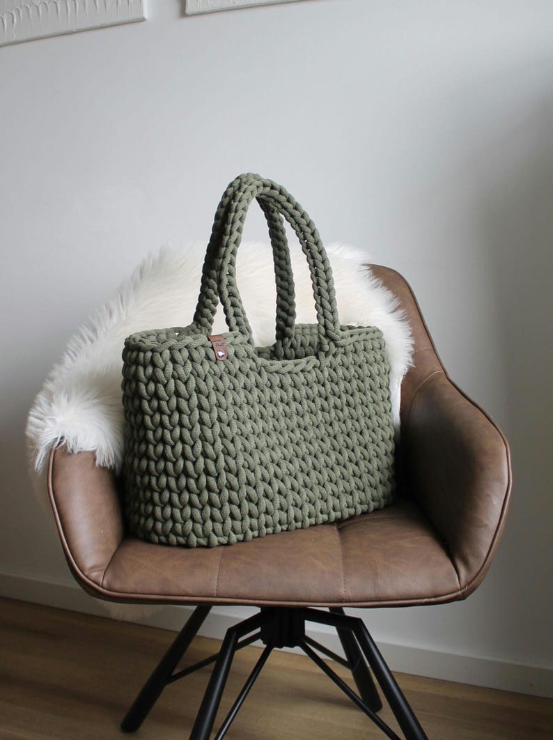 May include: A green crocheted tote bag with brown leather handles sits on a brown leather chair with a white faux fur cushion.