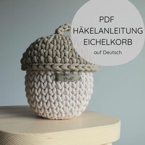 Op de afbeelding: Een gehaakte mand in de vorm van een eikel met een beige dop en een lichtcrème basis. De mand heeft een klein steeltje aan de bovenkant en een rechthoekig label met de tekst KNOTEN Punkt. De tekst op de afbeelding luidt PDF HÄKELANLEITUNG EICHELKORB auf Deutsch.