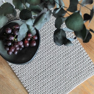Könnte beinhalten: Ein gewebtes Platzset in Grau und Weiß mit einem Lederanhänger. Das Platzset liegt auf einem Holztisch mit einer Schale Trauben und einem Zweig Eukalyptus.