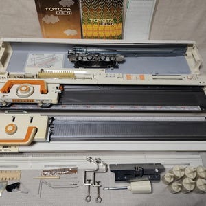 Peut inclure: Machine à tricoter Toyota KS901 vintage, ouverte et prête à l'emploi, avec des accents orange et blancs. L'image comprend les composants de la machine, les manuels d'instructions et les accessoires. La machine est conçue pour créer des textiles tricotés.