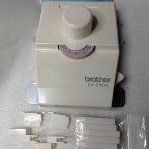 May include: A white Brother KA-2600 Intarsia Sled with a dial that reads 0, 1, 2, 3, and 4. The sled is accompanied by several white plastic pieces.