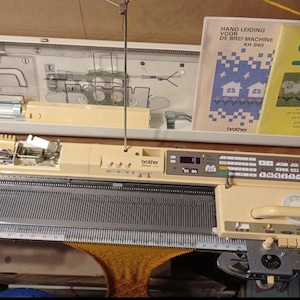 May include: A beige Brother KH 840 knitting machine with a digital display and a beige carrying case. The machine is accompanied by two books, one titled "Stitch World Pattern Book" and the other titled "Handleiding voor de Brei Machine KH 840".
