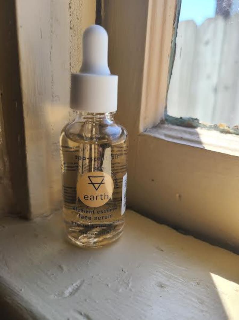 K&ouml;nnte beinhalten: Eine klare Glasflasche mit Gesichtsserum und einem wei&szlig;en Tropferaufsatz. Die Flasche hat ein rundes, beiges Etikett mit dem Wort "earth" und einem Dreieckssymbol. Das Serum hat eine hellbernsteinfarbene Farbe.