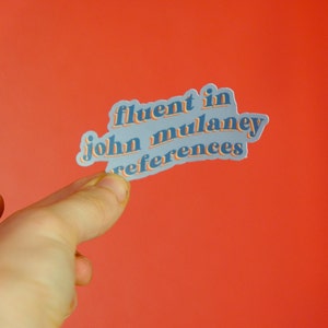 May include: A sticker with blue text on a white background that reads "fluent in john mulaney references".