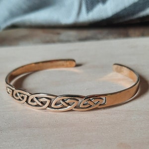 Könnte beinhalten: Ein goldfarbenes Armband mit keltischem Knotenmuster. Das Armband besteht aus glattem Metall und zeigt ein sich wiederholendes Muster aus ineinander verschlungenen Knoten.