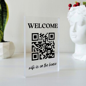 May include: A clear acrylic sign with the word "WELCOME" printed in black. A black and white QR code is below the word "WELCOME". The text "wifi is on the house" is printed in black below the QR code.