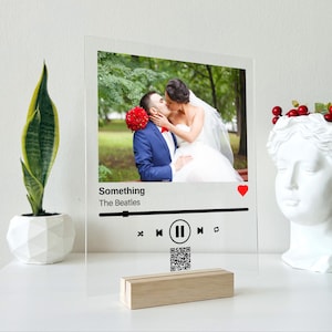 May include: A clear acrylic plaque featuring a printed photograph of a couple kissing, with the text Something and The Beatles, a red heart icon, a playback progress bar, media control icons, and a QR code, supported by a light wood base.